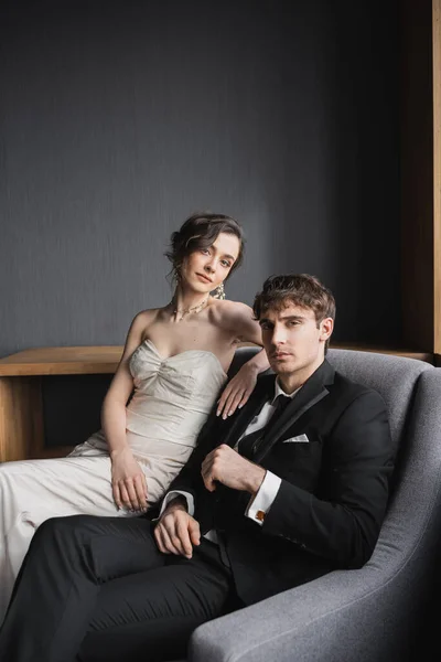 Брюнетка в білій весільній сукні і розкішні прикраси, що сидять на зручному кріслі разом з гарним виглядом нареченого в чорному костюмі в готельному номері — стокове фото
