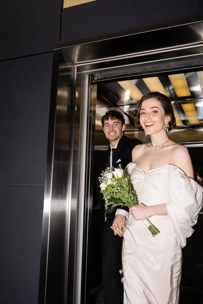 Bonita novia en vestido blanco sosteniendo ramo de novia con flores y mano de novio alegre en traje caminando fuera del ascensor en el hotel, recién casados felices - foto de stock