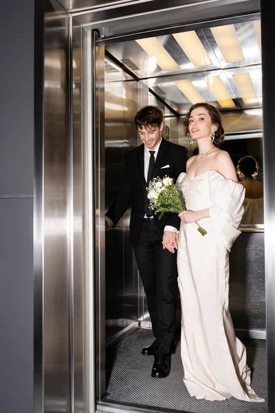 Full length of pretty bride in white dress holding bridal bouquet with flowers and hand of cheerful groom in suit standing in elevator in hotel, happy newlyweds — Stock Photo