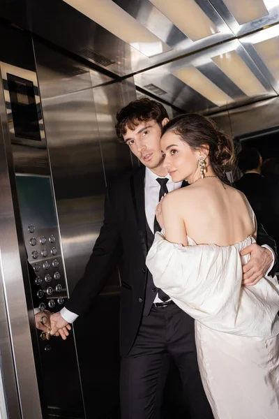 Gut aussehender Bräutigam in formeller Kleidung drückt Taste, während brünette Braut im Hochzeitskleid umarmt und zusammen im Fahrstuhl eines modernen Hotels steht — Stockfoto