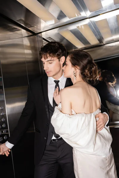 Good looking groom in formal wear pushing button while hugging brunette and young bride in wedding dress and standing together in elevator of modern hotel — Stock Photo