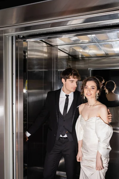 Гарний вигляд нареченого в офіційному одязі натискаючи кнопку і обіймаючи веселу наречену в весільній сукні, стоячи разом в ліфті сучасного готелю — стокове фото