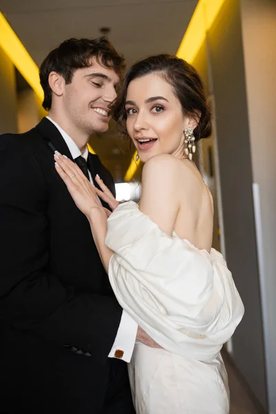 Noivo feliz em terno preto abraçando noiva animada em vestido de noiva branco e brincos de pérola enquanto sorrindo juntos no corredor do hotel moderno, casal na lua de mel — Fotografia de Stock