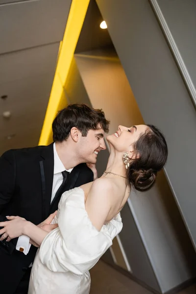 Side view of cheerful groom in black suit leaning towards charming bride in white wedding dress while smiling together in corridor of modern hotel, couple on honeymoon — Stock Photo