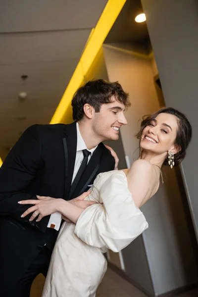 Noivo alegre em terno preto inclinando-se para a noiva encantadora em vestido de noiva branco, enquanto sorrindo juntos e de pé no salão do hotel moderno, casal na lua de mel — Fotografia de Stock
