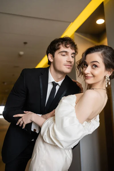 Весёлый мужчина в черном костюме склоняется к великолепной невесте в белом свадебном платье, улыбаясь вместе и стоя в коридоре современного отеля, концепция медового месяца — стоковое фото