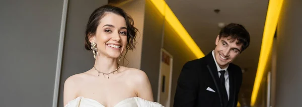 Hombre borroso y alegre en traje negro mirando a la novia hermosa en vestido de novia blanco mientras sonríen y caminan juntos en el pasillo del hotel moderno, bandera - foto de stock