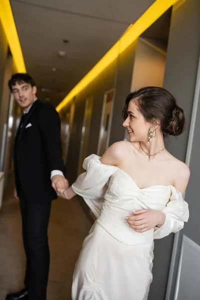 Gorgeous bride in white wedding dress holding hands with blurred and cheerful groom in black suit while smiling and walking together in corridor of modern hotel — Stock Photo
