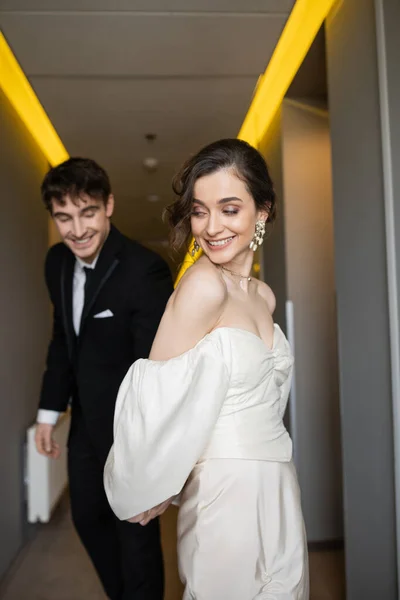 Восхитительная невеста в белом свадебном платье держится за руки с размытым и веселым женихом в черном костюме, улыбаясь и гуляя вместе в коридоре современного отеля, счастливых молодоженов в медовый месяц — стоковое фото