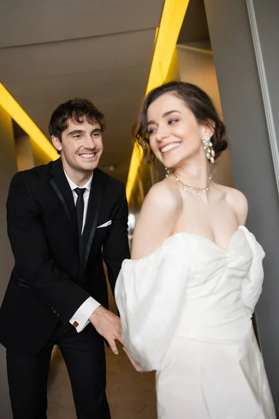 Cheerful groom in black suit tickling delightful bride in white wedding dress while smiling and walking together in hallway of modern hotel, happy newlyweds on honeymoon — Stock Photo