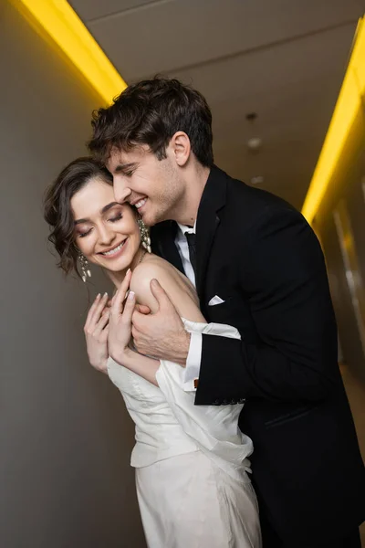 Cheerful groom in black suit embracing delightful bride in white wedding dress while smiling and standing together in hallway of modern hotel, happy newlyweds on honeymoon — Stock Photo