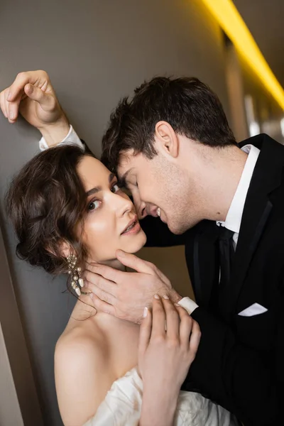 Smiling groom in black suit leaning towards wall and touching face of stunning bride in white wedding dress while standing together in hallway of modern hotel — Stock Photo