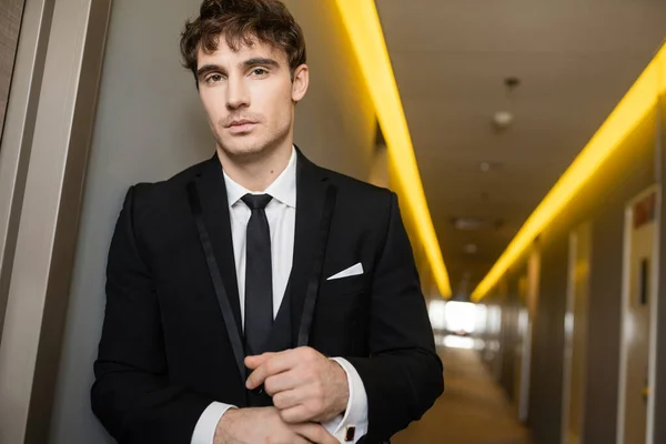 Portrait of good looking young groom in black suit with while shirt and tie looking at camera while standing in corridor of modern hotel on wedding day — Stock Photo