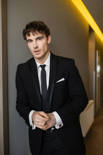 Portrait of handsome and young groom in black suit with while shirt and tie looking at camera while standing with opened mouth in corridor of modern hotel on wedding day — Stock Photo