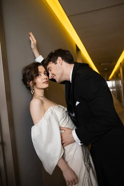 Cheerful groom in black suit leaning towards wall and hugging stunning bride in white wedding dress while standing together in hallway of modern hotel, newlyweds on honeymoon — Stock Photo