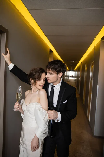 Молодий наречений в чорному костюмі спирається на стіну і тримає пляшку біля приголомшливої нареченої зі склянкою шампанського, стоячи разом в коридорі сучасного готелю, молодята на медовий місяць — стокове фото