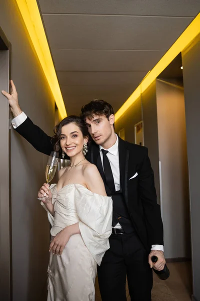 Good looking groom in black formal wear leaning towards wall and holding bottle near happy bride with glass of champagne while standing together in hallway of modern hotel — Stock Photo