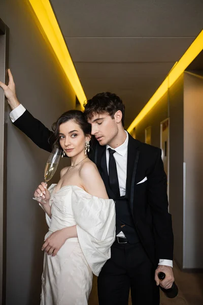 Groom in black suit leaning towards wall and holding bottle near charming bride with glass of champagne while standing together in corridor of modern hotel — Stock Photo