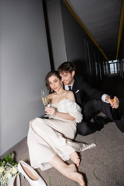 Noivo feliz em preto terno segurando garrafa e sentado perto de noiva encantadora com copo de champanhe ao lado de buquê de noiva e saltos altos no chão no corredor do hotel moderno — Fotografia de Stock