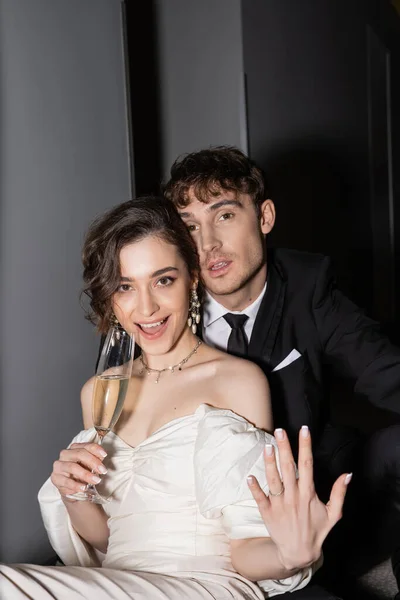 Взволнованная невеста в белом платье показывает руку с обручальным кольцом и держит бокал шампанского возле жениха с открытым ртом, глядя на камеру в холле отеля — стоковое фото