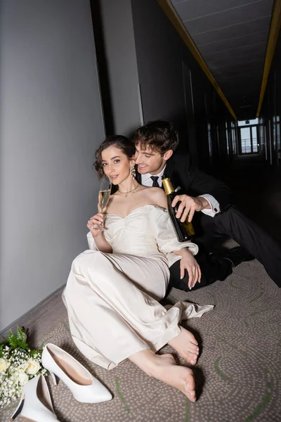 Happy groom in black suit holding bottle and looking at gorgeous bride drinking champagne next to bridal bouquet and high heels on floor in hall of modern hotel — Stock Photo