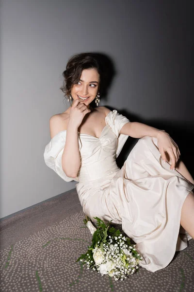 Happy young bride with brunette hair in white wedding dress smiling while looking away and sitting next to bridal bouquet with flowers on floor of hall in modern hotel — Stock Photo