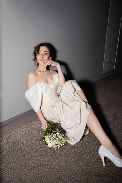 Young bride with brunette hair in white wedding dress smiling and looking at camera while sitting next to bridal bouquet with flowers on floor of hall in modern hotel — Stock Photo