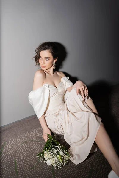 Radiant young bride with brunette hair in white wedding dress looking away while holding bridal bouquet with flowers and sitting on floor of hall in modern hotel — Stock Photo