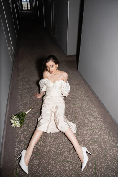 High angle view of young bride in white wedding dress and high heels looking at camera and sitting next to bridal bouquet with flowers on floor of hall in modern hotel — Stock Photo