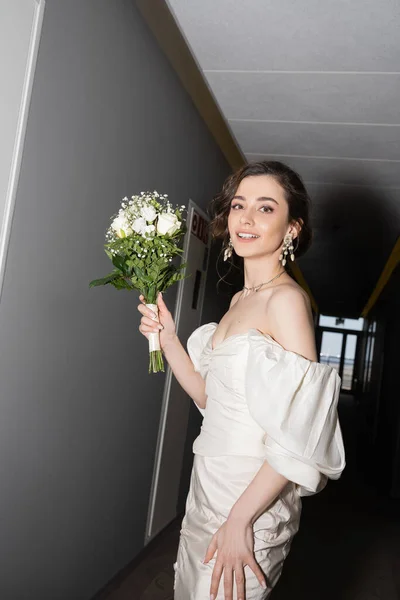 Cheerful and brunette young bride in white dress smiling while holding bridal bouquet with flowers and looking at camera in hall in modern hotel, beautiful bride on wedding day — Stock Photo