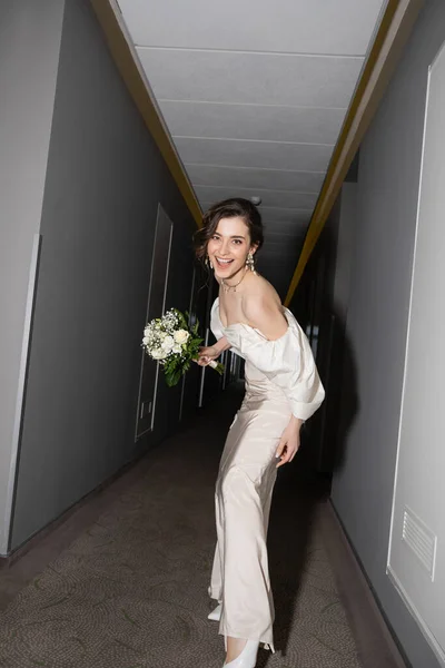 Aufgeregte und brünette junge Braut im weißen Brautkleid lächelnd, während sie den Brautstrauß mit Blumen in der Hand hält und in die Kamera im Saal des modernen Hotels blickt, wunderschöne Braut — Stockfoto