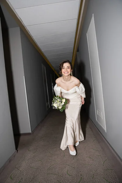 Full length of excited and brunette young bride in white wedding dress smiling while holding bridal bouquet with flowers and looking at camera in hall in modern hotel — Stock Photo