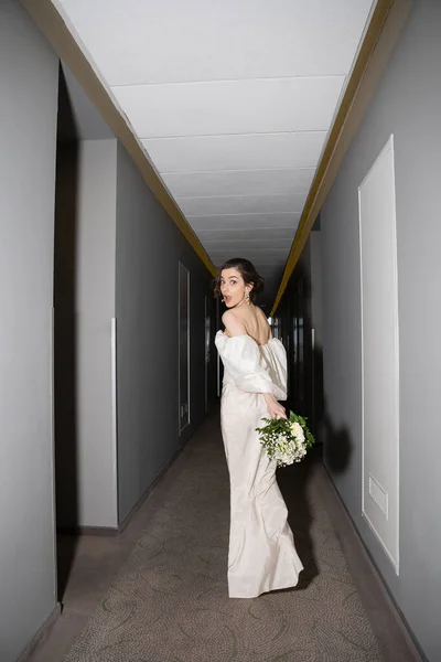 Full length of stunned and brunette bride with opened mouth standing in white wedding dress and holding bridal bouquet with flowers while looking at camera in hall in modern hotel — Stock Photo