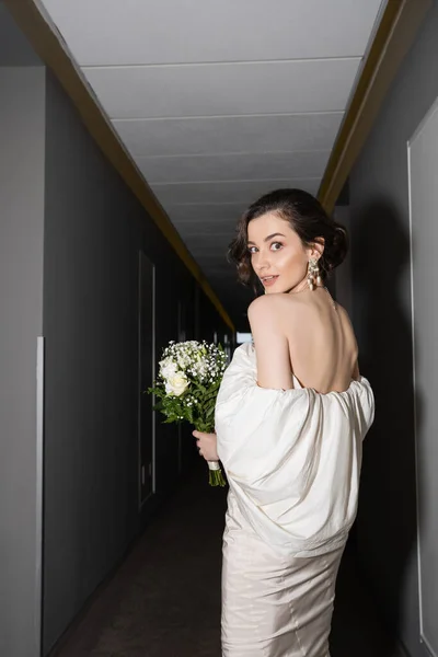 Fassungslos und brünett Braut in weißem Brautkleid hält Brautstrauß mit Blumen und blickt in die Kamera, während sie im Flur des modernen Hotels stehen — Stockfoto