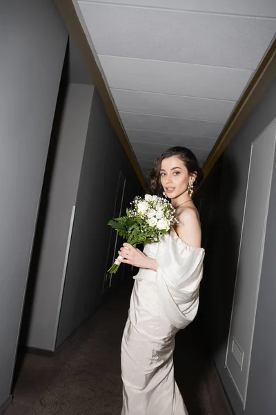 Young and brunette bride in white wedding dress holding bridal bouquet with flowers and looking at camera while standing in hallway of modern hotel — Stock Photo