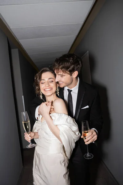 Joven y alegre novia en vestido de novia blanco y novio alegre sosteniendo copas de champán mientras están de pie y sonriendo juntos en el pasillo del hotel, recién casados en luna de miel - foto de stock
