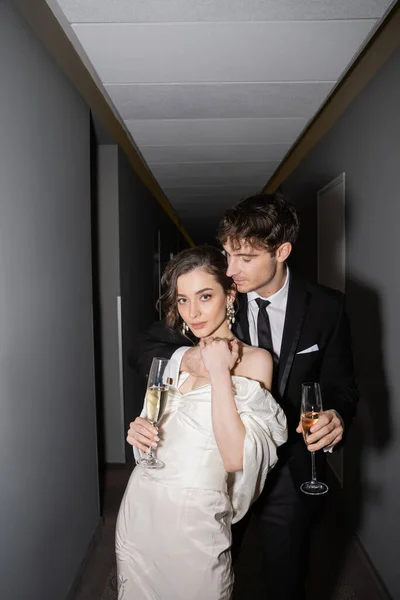 Young and brunette bride in white wedding dress and handsome groom holding glasses of champagne while standing together in hallway of hotel, newlyweds on honeymoon — Stock Photo