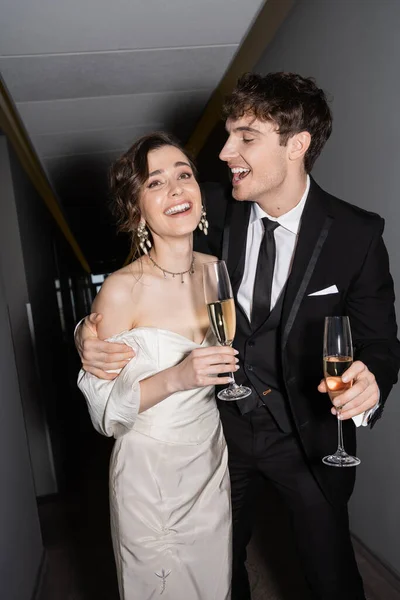 Cheerful groom hugging young and brunette bride in white wedding dress and holding glasses of champagne while standing and smiling together in hallway of hotel, newlyweds on honeymoon — Stock Photo