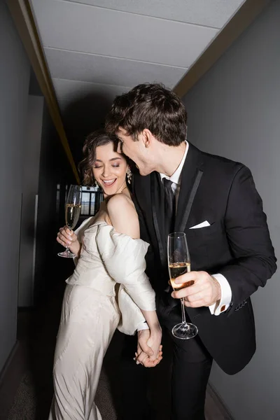 Excited groom hugging young and brunette bride in white wedding dress and holding glasses of champagne while standing and smiling together in hallway of hotel — Stock Photo