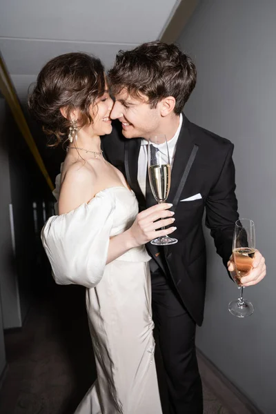 Cheerful groom hugging and kissing young and brunette bride in white wedding dress and holding glasses of champagne while smiling together in hallway of hotel, newlyweds on honeymoon — Stock Photo