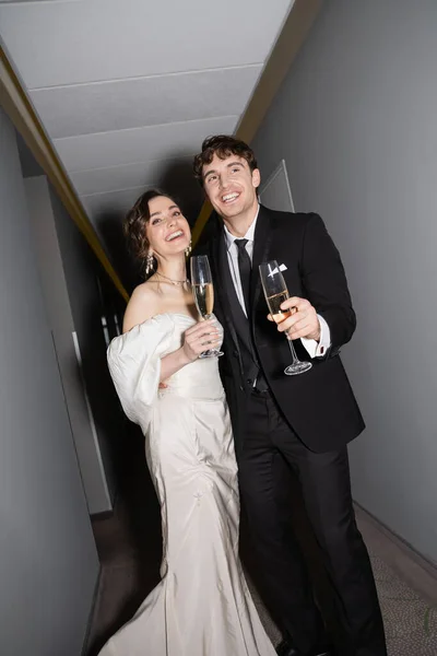 Cheerful groom and young bride in white wedding dress and holding glasses of champagne while standing and smiling together in hallway of hotel, newlyweds on honeymoon — Stock Photo