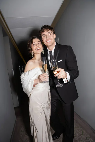 Noivo alegre abraçando noiva jovem e morena em vestido de noiva branco e segurando copos de champanhe enquanto de pé e sorrindo juntos no corredor do hotel — Fotografia de Stock