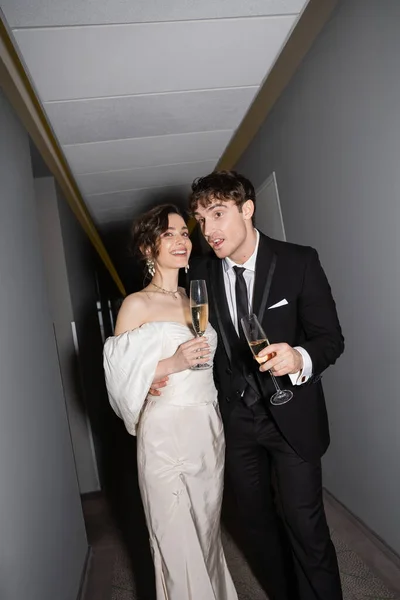 Sposo emotivo abbracciare giovane e felice sposa in abito da sposa bianco e tenere i calici di champagne mentre in piedi e sorridente insieme nel corridoio dell'hotel — Foto stock