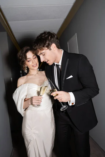 Cheerful groom and brunette bride in white wedding dress and clinking glasses of champagne while standing and smiling together in hallway of hotel, newlyweds on honeymoon — Stock Photo