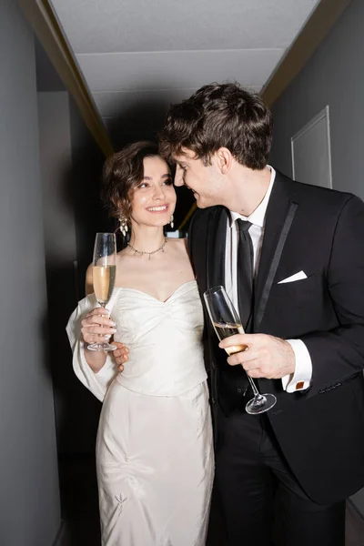 Noivo alegre abraçando noiva jovem e feliz em vestido de casamento branco e segurando copos de champanhe enquanto de pé e sorrindo juntos no corredor do hotel, recém-casados na lua de mel — Fotografia de Stock
