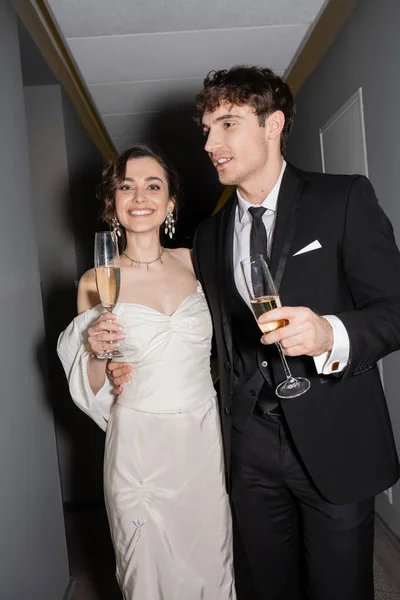 Handsome groom hugging young and happy bride in white dress and holding glasses of champagne while standing and smiling together in corridor of hotel — Stock Photo