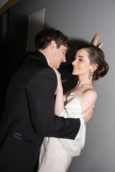 Счастливый жених в черном костюме склоняется к стене и смотрит на невесту в белом свадебном платье, улыбаясь и стоя вместе в коридоре современного отеля, молодожены в медовый месяц — стоковое фото