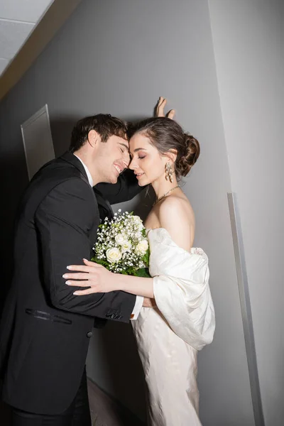 Позитивный жених в черном костюме склоняется к стене рядом с невестой в белом свадебном платье держа свадебный букет, стоя вместе в коридоре современного отеля, молодожены в медовый месяц — стоковое фото