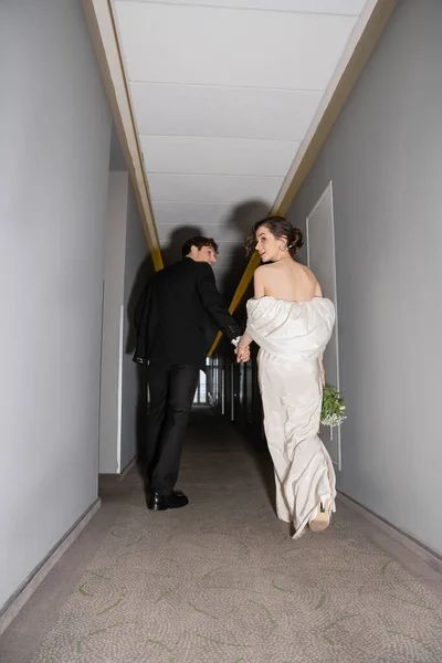 Low angle view of groom in black suit holding hands with bride in white wedding dress carrying bridal bouquet while walking together in hallway of modern hotel, newlyweds on honeymoon — Stock Photo