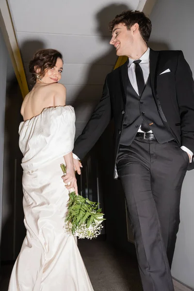 Низкий угол зрения веселого жениха в черном костюме позируя с рукой в кармане и глядя на счастливую невесту в белом свадебном платье держа свадебный букет во время прогулки вместе в зале современного отеля — стоковое фото
